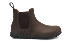 A brown leather Chelsea Boot by Xero Shoes featuring a black rubber sole, elastic side panel, pull tab at the back, and minimal stitching, depicted from the side on a white background—ideal for those seeking a classic style and fit guide.