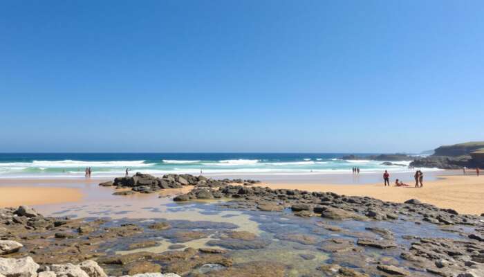 A sunlit beach with clear waves, golden sands for sunbathing, rocky outcrops for exploration, accessible ramps, and diverse people enjoying the day under a blue sky.